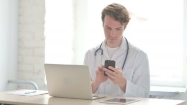 Male Doctor using Smartphone while using Laptop in Clinic 