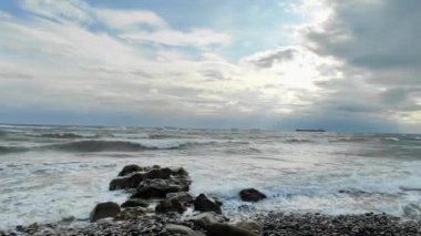Deniz bulutlu bir günde kıyıda kayalara çarpıyor ve arka planda bir gemi var.