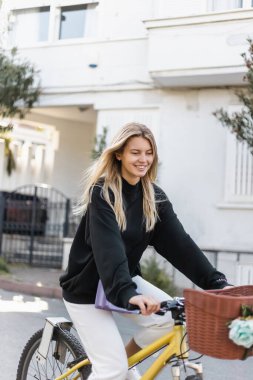 Moda kıyafetli neşeli genç bir kadın hindiyle sokakta bisiklet sürüyor.