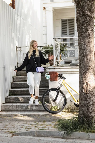 young and cheerful woman in trendy outfit walking on stairs near house and bicycle