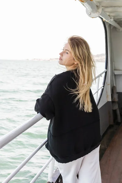 Mujer rubia joven en suéter negro mirando el mar desde el ferry que cruza el estrecho del Bósforo en Estambul - foto de stock