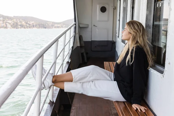 Femme blonde en pull noir assis sur le banc tout en regardant la mer de ferry bateau traversant le bosphore à Istanbul — Stock Photo