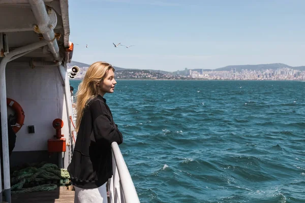 Вид сбоку счастливой женщины в черном свитере, смотрящей в море с паромной переправы в Стамбуле — стоковое фото