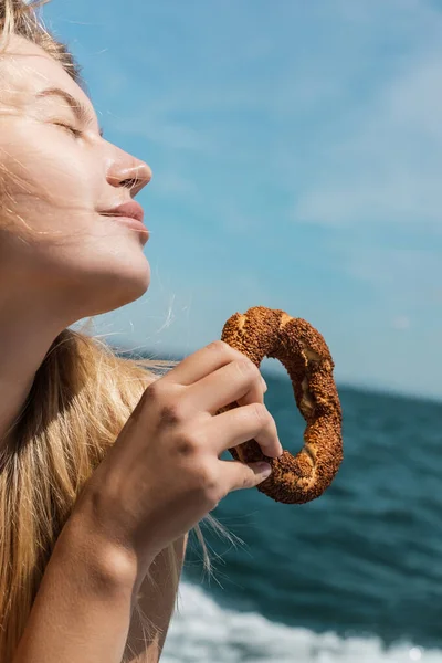 Pleased blonde woman with closed eyes holding turkish sesame bagel near sea — Stock Photo