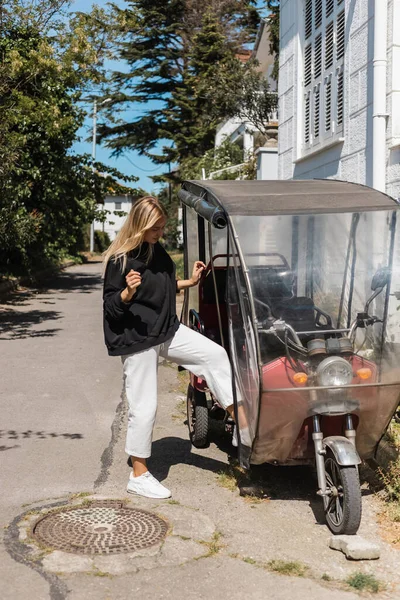 Jeune femme blonde en tenue élégante près de cyclomoteur à trois roues avec toit en dinde — Photo de stock