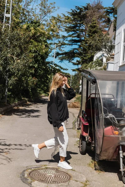 Mujer rubia en traje elegante caminando cerca de tres ruedas ciclomotor con techo en pavo - foto de stock