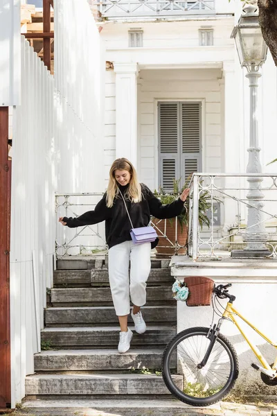 Jeune et heureuse femme en tenue tendance marchant sur les escaliers près de la maison et du vélo — Photo de stock