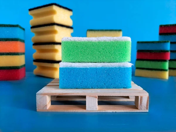 Two kitchen sponges on wooden pallet. Blurred background. In foreground, washcloths for washing dishes lie on stand. In background are stacks of sponges for kitchen. Concept of choosing right cleaning accessories