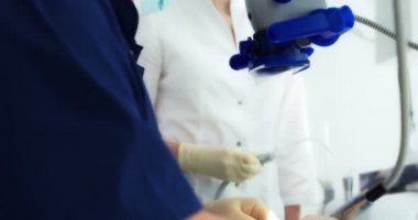 Maskeli bir dişçi ve eldiven giymiş bir asistan modern bir diş muayenehanesindeki bir hastaya mikroskop altında profesyonel diş temizliği yapıyorlar..