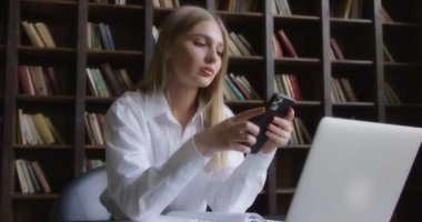 Beyaz gömlekli bir iş kadını dizüstü bilgisayarın önünde telefonla konuşuyor.