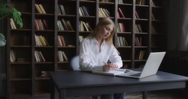 Liikenainen valkoisessa paidassa työskentelee kannettavassa tietokoneessa ja tekee muistiinpanoja. — kuvapankkivideo