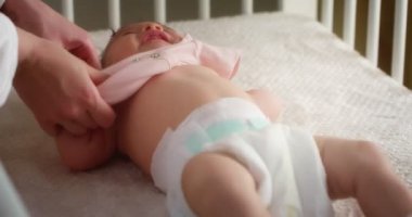 Annem bir bebek beşiğinde bebek bezinde yatan bir bebeğe sevgiyle pembe bir beden giydirir.