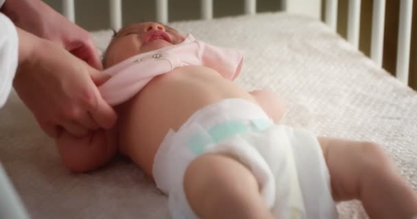 Mom lovingly puts a pink bodysuit on a newborn lying in diaper in a child crib — Stockvideo