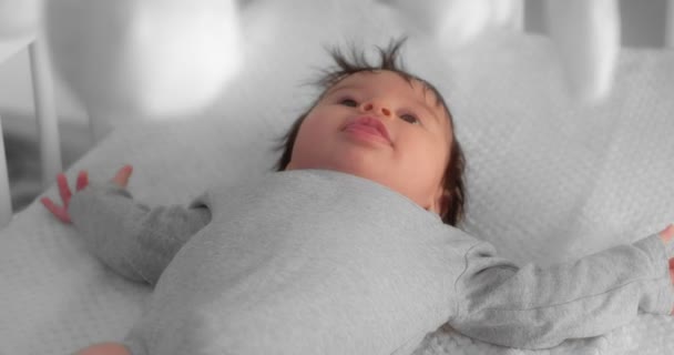 Bebé recién nacido en un traje gris está acostado en una cuna y mirando a un niño móvil — Vídeos de Stock
