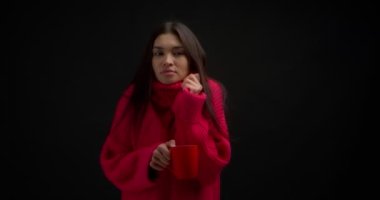 Pembe kazaklı, uzun saçlı güzel esmer donmuştu. Asyalı bir kadın soğuk algınlığı geçirir, kırmızı bardakta sıcak bir içecekle ısınır. Siyah bir arkaplanda izole edilmiş.