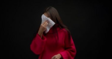 Pembe kazaklı hasta Asyalı kadın ağzını kağıt peçeteyle kapatıyor ve termometreye bakıyor. Siyah arka planda izole edilmiş genç bir kadında burun akıntısı, ateş ve öksürük.. 