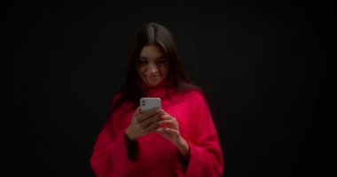 Parlak pembe kazaklı tatlı Asyalı kadın telefonunda hoş bir mesaj okuyor ve izole edilmiş siyah bir arka planda dururken kameraya rüya gibi gülümsüyor.. 