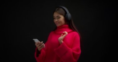 Parlak pembe kazaklı güzel bir kadın telefonunda müzik açıyor, en sevdiği şarkı kulaklıklarında çalıyor. Pozitif Asyalı kadın siyah arka planda müzik dinliyor..