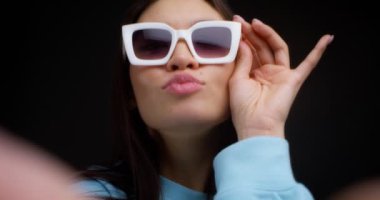 Beyaz gözlüklü, akıllı telefondan selfie çeken genç ve güzel bir esmerin yakın çekimi. Mavi elbiseli genç bir kadın siyah arka planda telefonuyla kendi fotoğrafını çekiyor.. 