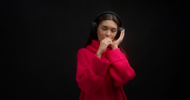 Woman listens to music in wireless headphones, smokes vape on a black background — Stock Video