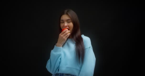Jovem bela morena em um jumper azul morde uma maçã vermelha. Alimentos saudáveis. — Vídeo de Stock