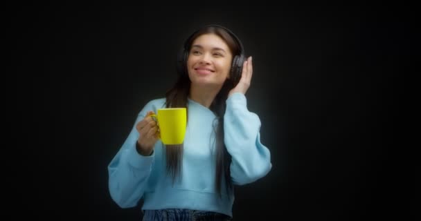 Mavi kazaklı kadın kulaklıklı müzikten hoşlanıyor, sarı bardaktan içiyor. — Stok video