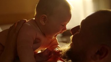 Sakallı, yeni doğmuş bebeğini okşayan ve öpen genç bir baba. Annesi tarafından sabah mutfakta kollarında tutuluyor. Aile kavramı, babalık, annelik, ebeveynlik, çocukluk, yaşam.