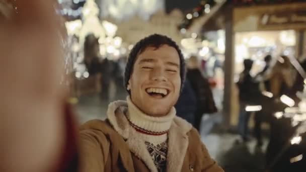Funny young man smiling and making selfie with his smartphone at amazing street fair in old European city on Christmas Eve. In the background ordinary people walking and buying souvenirs. — Stock Footage Funny young man smiling and making selfie with his smartphone at amazing street fair in old European city on Christmas Eve. In the background ordinary people walking and buying souvenirs. Stock Video