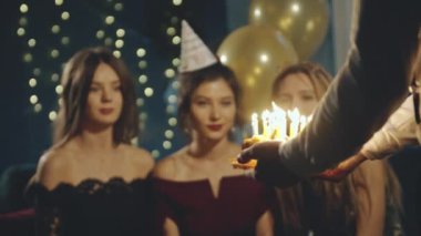 Genç bir kadın mumlara ateş üflüyor ve partilerde ya da kutlamalarda dilek tutuyor. Gülümseyen Asyalı kız ve arkadaşlarının yakın plan portresi. Sürpriz yapıyorlar ve mumlarla doğum günü pastasını çıkarıyorlar..