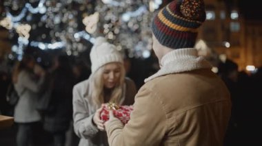 Kameraya karşı duran adam, eski kasaba meydanındaki Noel gecesinde kız arkadaşına bir hediye sunar. Aşk, aile, Noel, yeni yıl, tatil konsepti. RED kamera, 10 bit klonlama