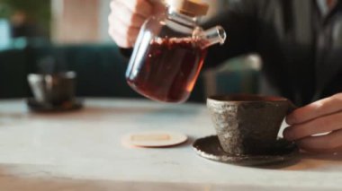 Kafe ziyaretçisinin çaydanlıktan güzel, orijinal çay fincanına çay dökerkenki yakın çekimi. Aromatik sıcak içecek. Meyveler havayı kokuyla dolduruyor. Restoranda ilginç kavramsal yemekler..