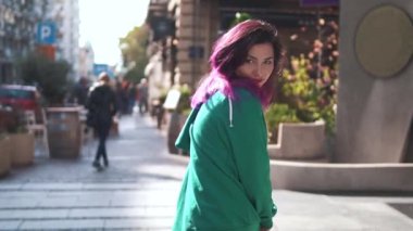 Şık, gülen kadın pozu. Gülümseyen kız kamerada flört ediyor. Güzel bir kadın eski şehir sokağına gidiyor ve güneş parlıyor. Mor saçlı, seksi ve şık bir kadının arka plan görüntüsü.