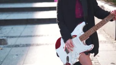 Elektro gitarlı genç rock kızı. Parlak günbatımında şehir merkezinde elektro gitar çalan mor saçlı cesur asi kadın. Arka planda yürüyen insanlar. Asyalı kız rock yıldızı gibi