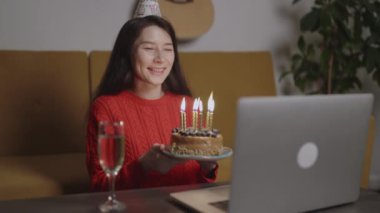 Doğum günü şapkalı mutlu Asyalı kadın tatildeki pastadaki mumları salgın covid-19 sırasında evde görüntülü sohbet yoluyla üflerken arkadaşını kutluyor. İnsanlar sosyal mesafeyi korumak için zaman harcar