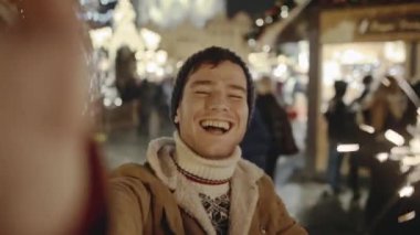 Eski Avrupa şehrinde Noel arifesinde muhteşem sokak fuarında gülümseyen ve akıllı telefonuyla selfie çeken komik genç adam. Arka planda sıradan insanlar yürüyor ve hediyelik eşya alıyor..