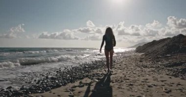 Tatlı kız kayalık sahilde yavaşça yürüyor. Plaj boyunca ilerleyen siyah mayolu çekici bir kadın. Deniz kenarındaki esmerin sinematik çekimi. Deniz dalgaları kızların ayaklarına vuruyor. Sanat evi kavramı