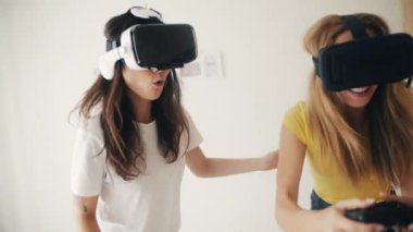 Evde sanal gerçeklik kasklı kızlar var. Sanal gerçeklik kulaklığı takmış iki kadın yatakta oturmuş, video oyunları oynuyor ve birlikte eğleniyorlar. Üç boyutlu teknoloji. Sanal gerçeklik, oyun, arkadaşlık kavramı.