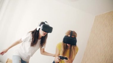 Evde sanal gerçeklik kasklı kızlar var. Sanal gerçeklik kulaklığı takmış iki kadın yatakta oturmuş, video oyunları oynuyor ve birlikte eğleniyorlar. Üç boyutlu teknoloji. Sanal gerçeklik, oyun, arkadaşlık kavramı.