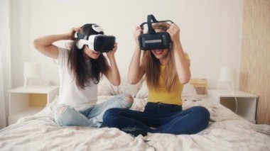 Evde sanal gerçeklik kasklı kızlar var. Sanal gerçeklik kulaklığı takmış iki kadın yatakta oturmuş, video oyunları oynuyor ve birlikte eğleniyorlar. Üç boyutlu teknoloji. Sanal gerçeklik, oyun, arkadaşlık kavramı.