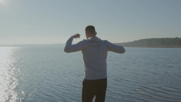 El hombre está haciendo rotaciones de brazos en la orilla del lago — Vídeos de Stock