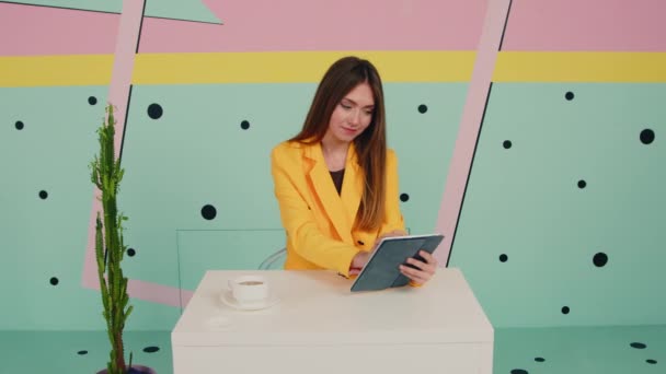 Mulher em casaco amarelo usa computador tablet e olha para a câmera — Vídeo de Stock