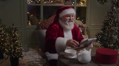 Noel Baba rahat arka bahçede oturuyor, ışıklarla süslenmiş, işiyle meşgul, çocuklardan gelen mektupları açıyor, Noel geliyor, Ağır çekim.