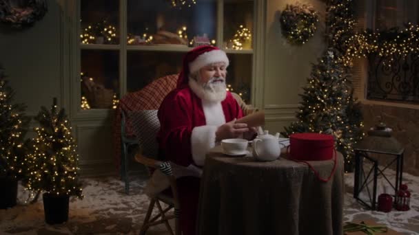 サンタクロースは彼の家の居心地の良い裏庭に座っています 照明やクリスマスツリーで飾られ お願いで子供たちからの手紙を読んでいます — ストック動画