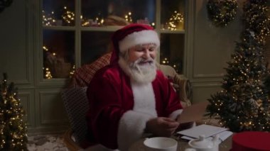 Noel Baba evinde oturuyor, ışıklarla ve Noel ağaçlarıyla süslenmiş, çocuklardan mektuplar okuyor ve gülümsüyor, mutlu Noeller diliyor, ön plan, yavaş çekim.