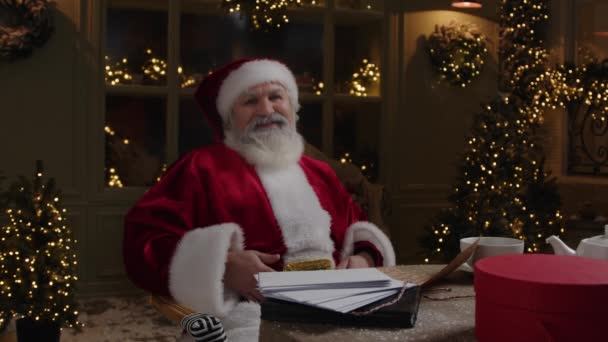 快乐的圣诞老人 一个留着灰白胡子的老人 坐在桌旁 对着摄像机笑着 分享节日的喜悦 圣诞节来了 慢动作 — 图库视频影像