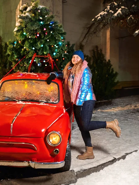 Atkı, şapka, ceket, kırmızı vintage araba ve dışarıda noel ağacı olan mutlu gülümseyen beyaz kadın. Yeni yıl, eğlence, kış konsepti