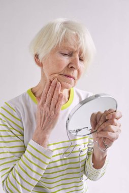 Yaşlı üzgün kadın aynada kendine bakıyor ve yüzüne dokunuyor. Yaş, zaman, cilt bakımı kavramı