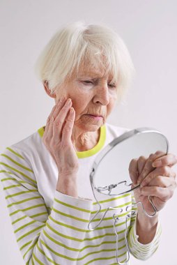 Yaşlı üzgün kadın aynada kendine bakıyor ve yüzüne dokunuyor. Yaş, zaman, cilt bakımı kavramı