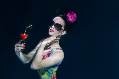 beautiful woman in a colourful stylish swimsuit and sunglasses and pink gerbera in the head with a cocktail  in her hand underwater in the swimming pool