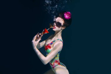 beautiful woman in a colourful stylish swimsuit and sunglasses and pink gerbera in the head with a cocktail  in her hand underwater in the swimming pool